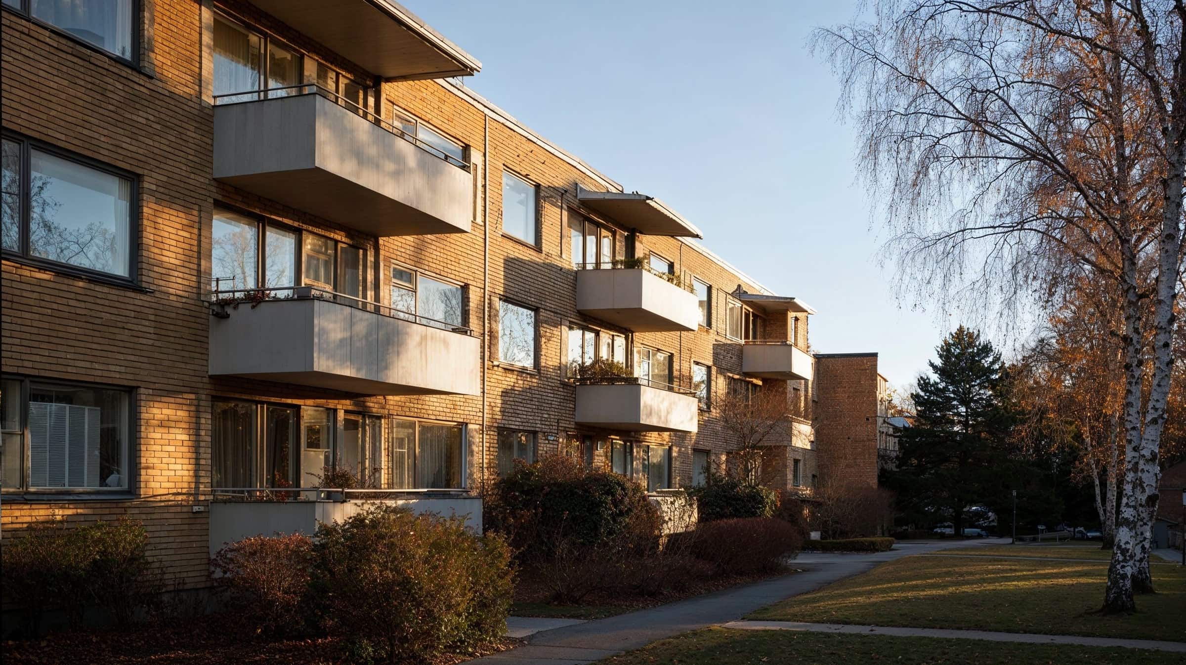Flerbostadshus i gult tegel med vita balkonger, klippta buskar längs fasaden och björkar på gården i låg eftermiddagssol.