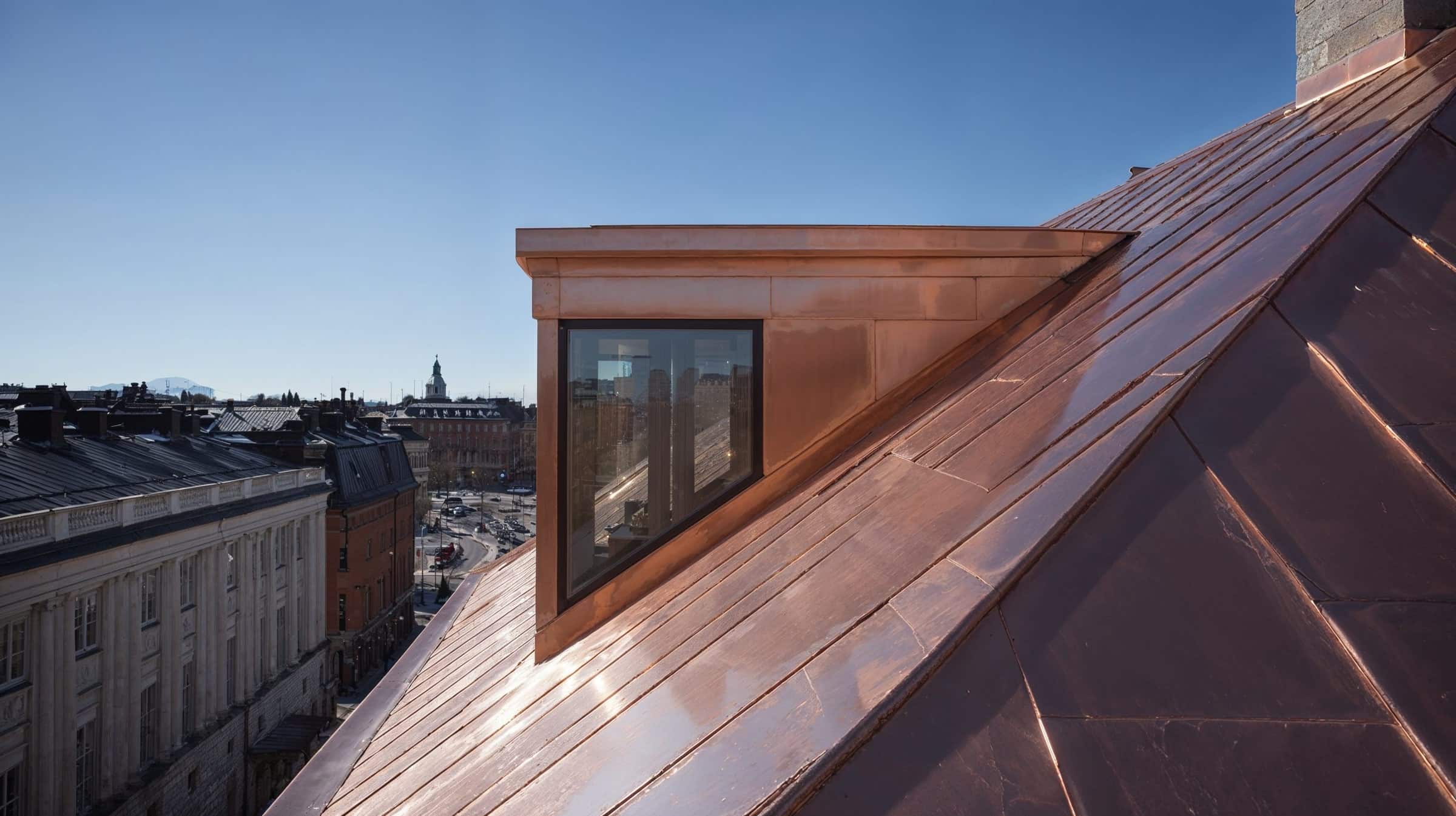Ett tak av blank, opatinerad koppar med en takkupa och ett stort fönster. I bakgrunden stadens tak och en kyrkspira.
